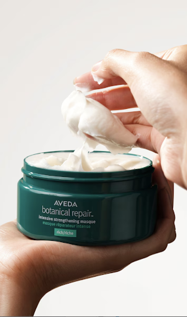 A person scooping out a creamy white product from an open jar labeled "Aveda Botanical Repair Intensive Strengthening Masque. - Scott J Salons in New York, NY